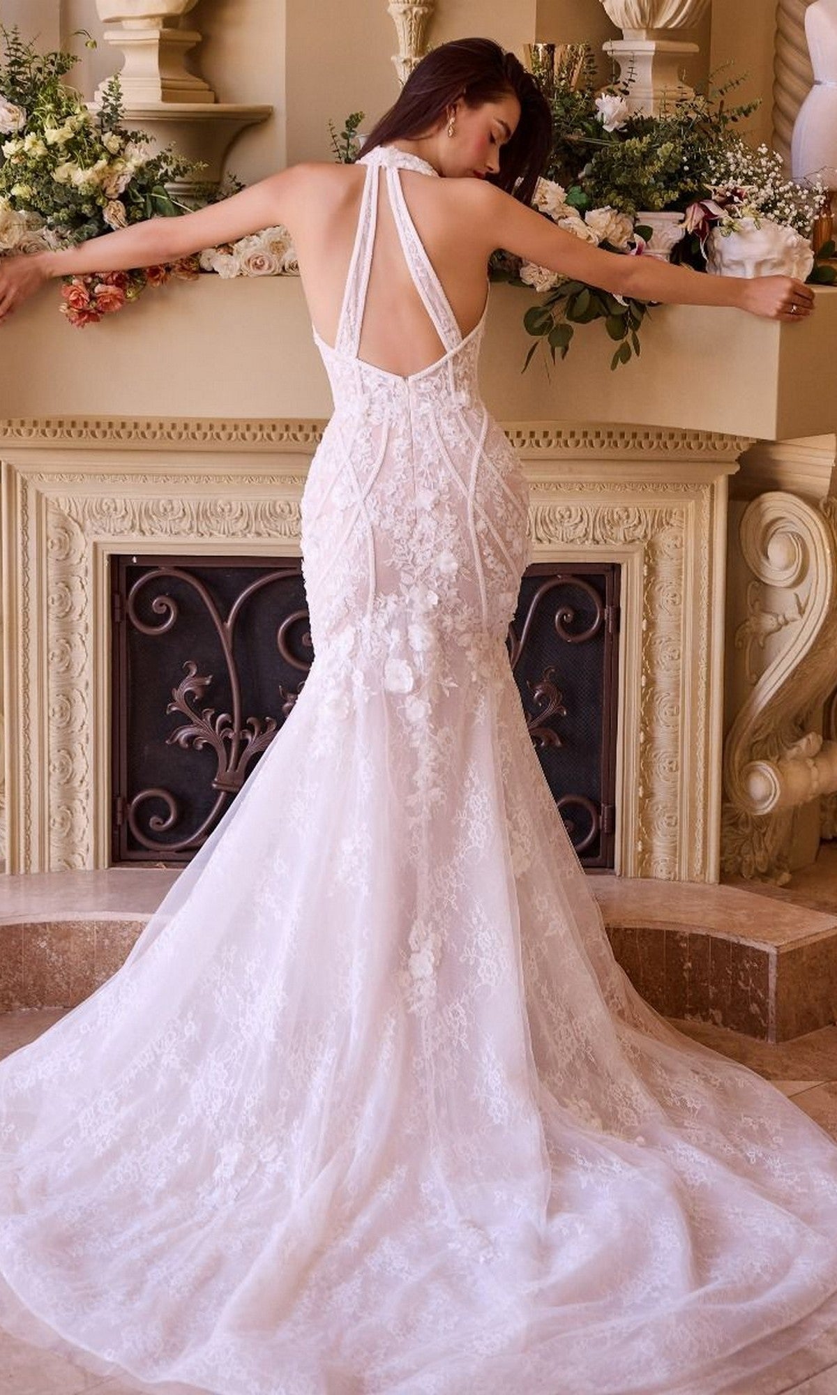 A woman wearing the Long Wedding Dress: Andrea and Leo WL019, featuring white lace, floral appliques, and a long train, stands facing a decorated fireplace with arms extended to either side.